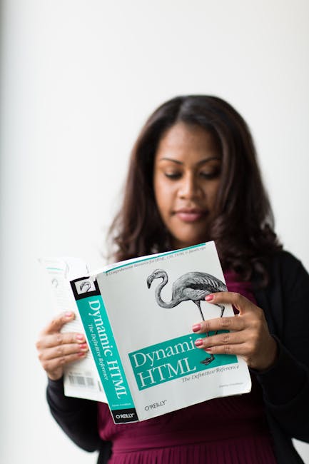 A woman with dark hair and a focused expression is holding a book titled 'Dynamic HTML' by O'Reilly, featuring a cover with a flamingo illustration. She is dressed in a black top and maroon cardigan, 